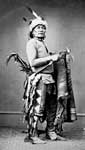 Dakota Indians: Portrait of Siha Han Ska (Long Foot) in Partial Native Dress with Headress and Ornaments and Holding Pipe Tomahawk - 1867